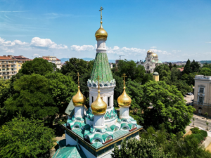 Руската църква в София – златните кубета и зеленият покрив, извисяващи се над зеленината в центъра на града. В далечината се вижда храмът „Св. Александър Невски